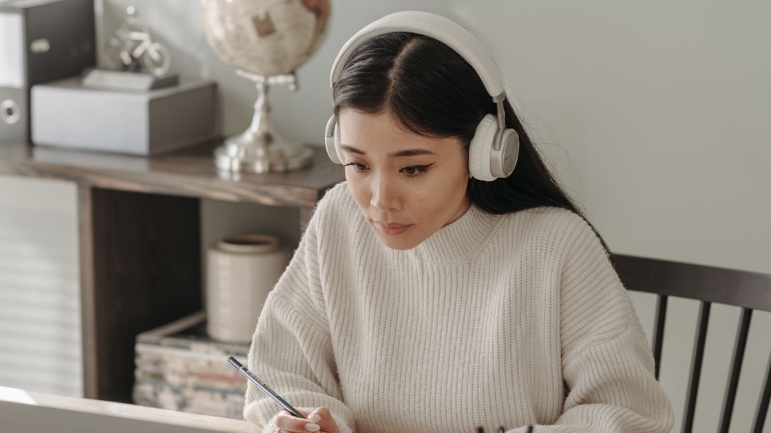 Uma estudante de fones revisa a aula com notebook e caderno em casa.